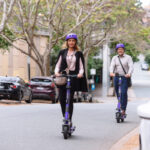 Two people wearing helmets riding on e-scooters