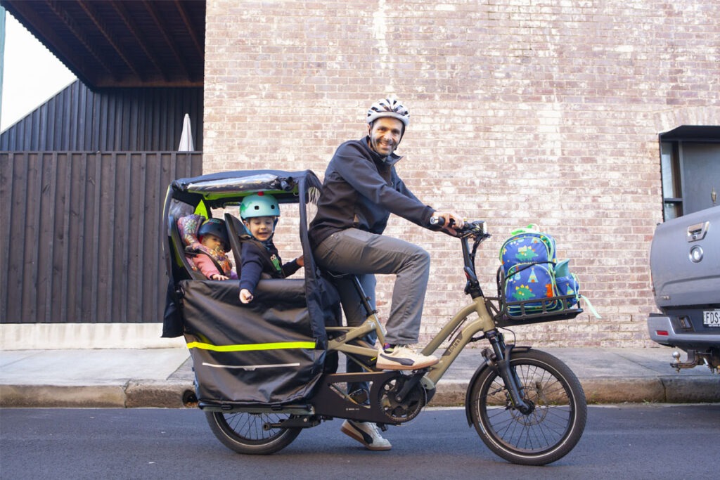 Man on e-cargo bike with two children seated in the back and backpack in the front.