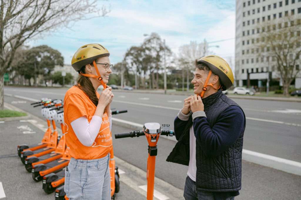 Two people putting on gold coloured helmets at e-scooter bay.