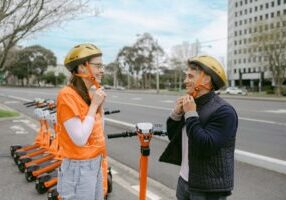 Two people putting on gold coloured helmets at e-scooter bay.